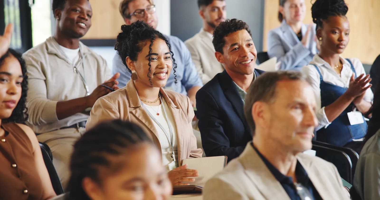 A diverse group of adults sitting in an audience, some smiling and clapping, while attending a conference or presentation.
