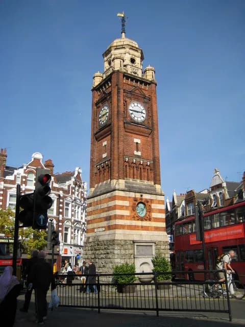 A towering brick clock tower with a dome and ornate details stands prominently in a city street, with buildings, traffic lights, and people visible around it.