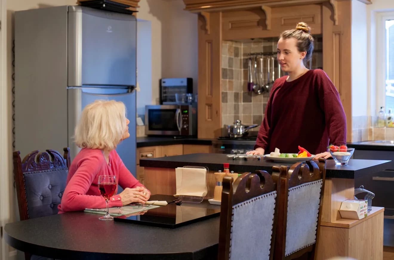A younger woman with hair in a bun stands at a kitchen island talking to an older woman with blonde hair sitting at a table in the foreground, with a glass of red wine in front of her.