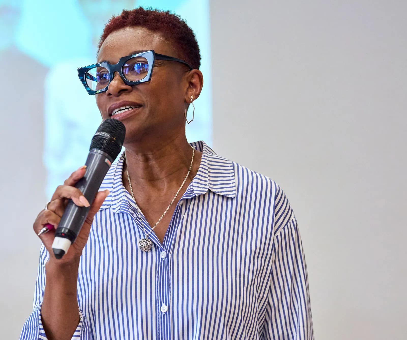 A woman with red hair and blue glasses speaks into a microphone while wearing a blue and white striped shirt.