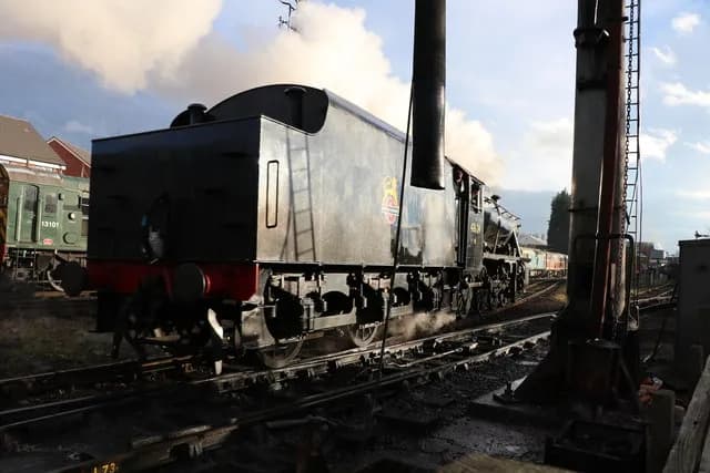 A large, dark steam locomotive with a tall smokestack and wisps of steam coming from it, sits on train tracks with other train cars visible in the background and signs to the left.