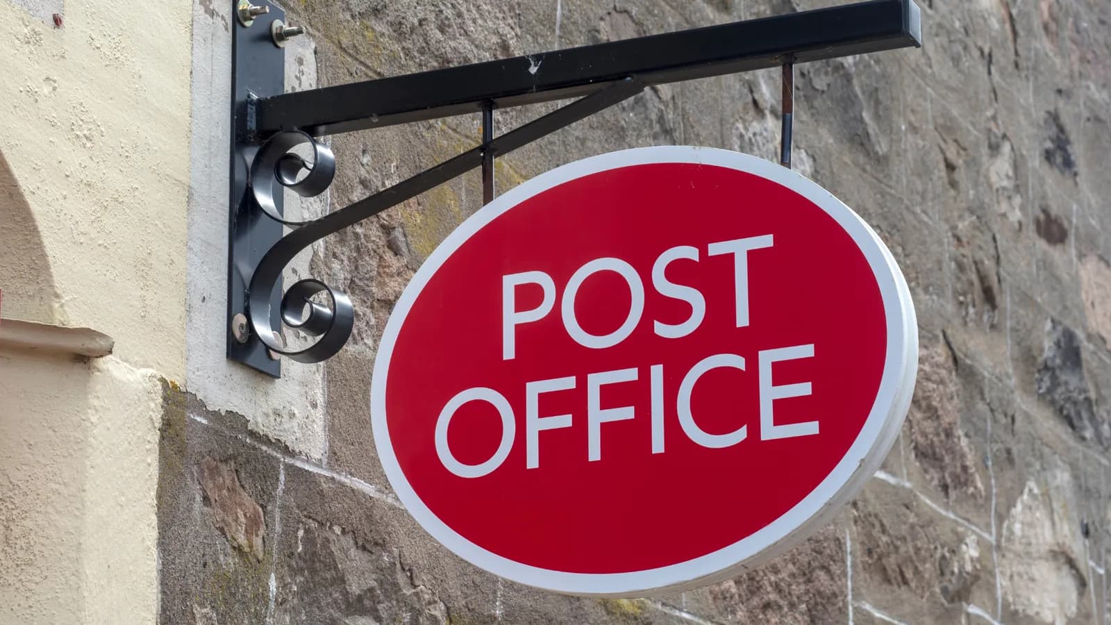 A bright red oval sign with white text reading "POST OFFICE" hangs from a black metal bracket against a stone and stucco building.