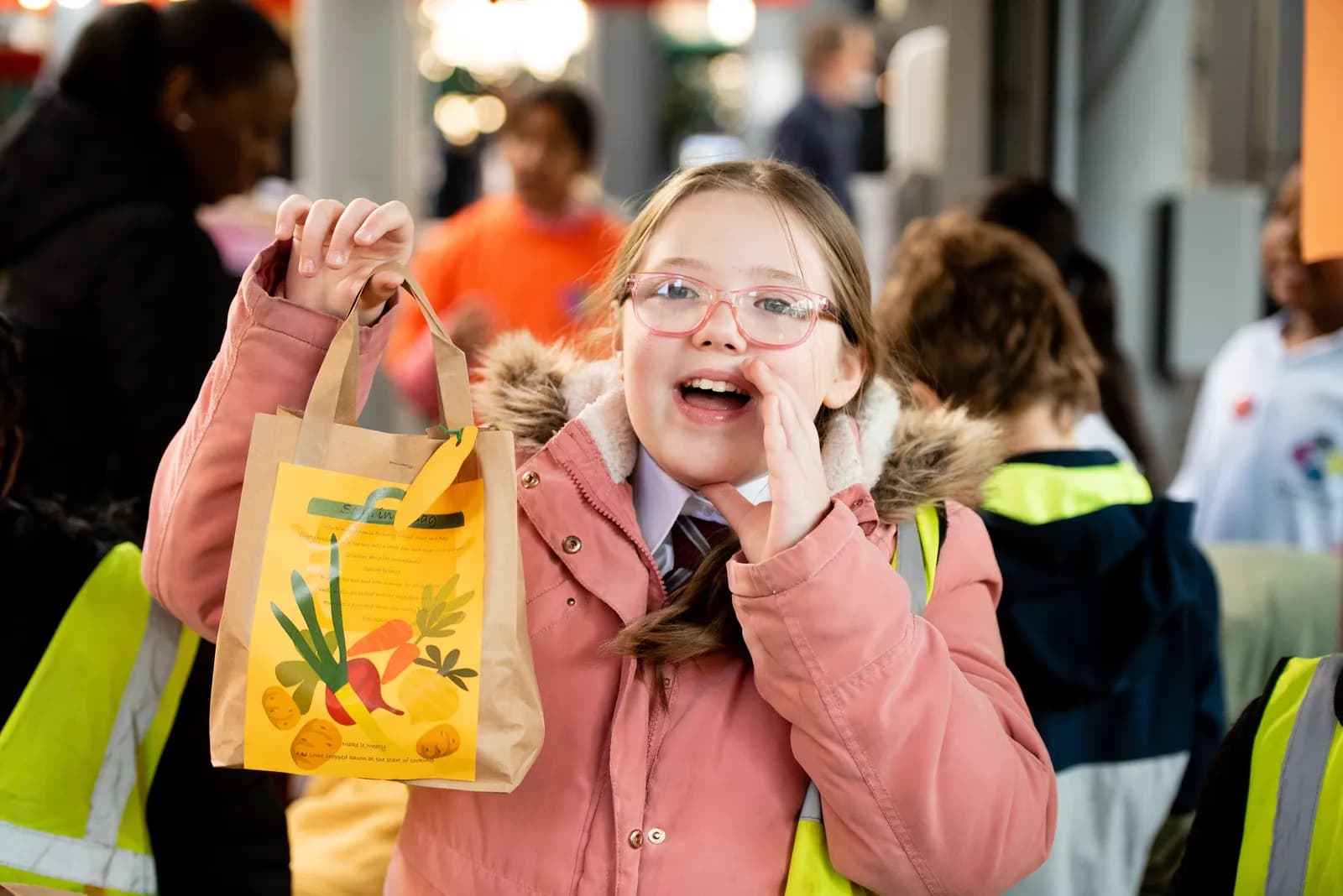 A young girl with glasses and a pink coat holds up a small paper bag with an illustration of vegetables, calling out to someone.