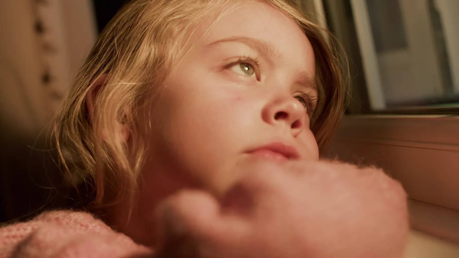 A young girl with blonde hair looks thoughtfully out of a window with her hand near her chin.