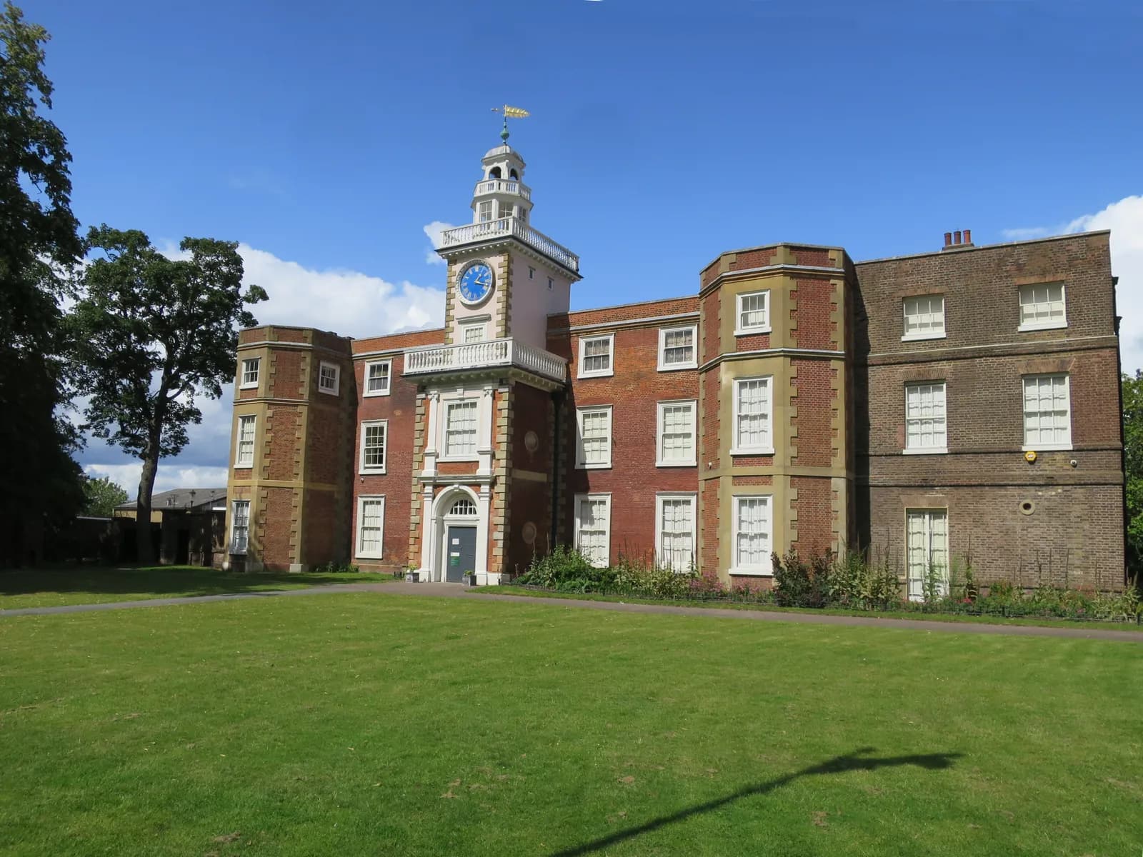 A historic brick mansion with many windows and a prominent central clock tower with a weather vane on top, surrounded by green grass and trees under a blue sky.