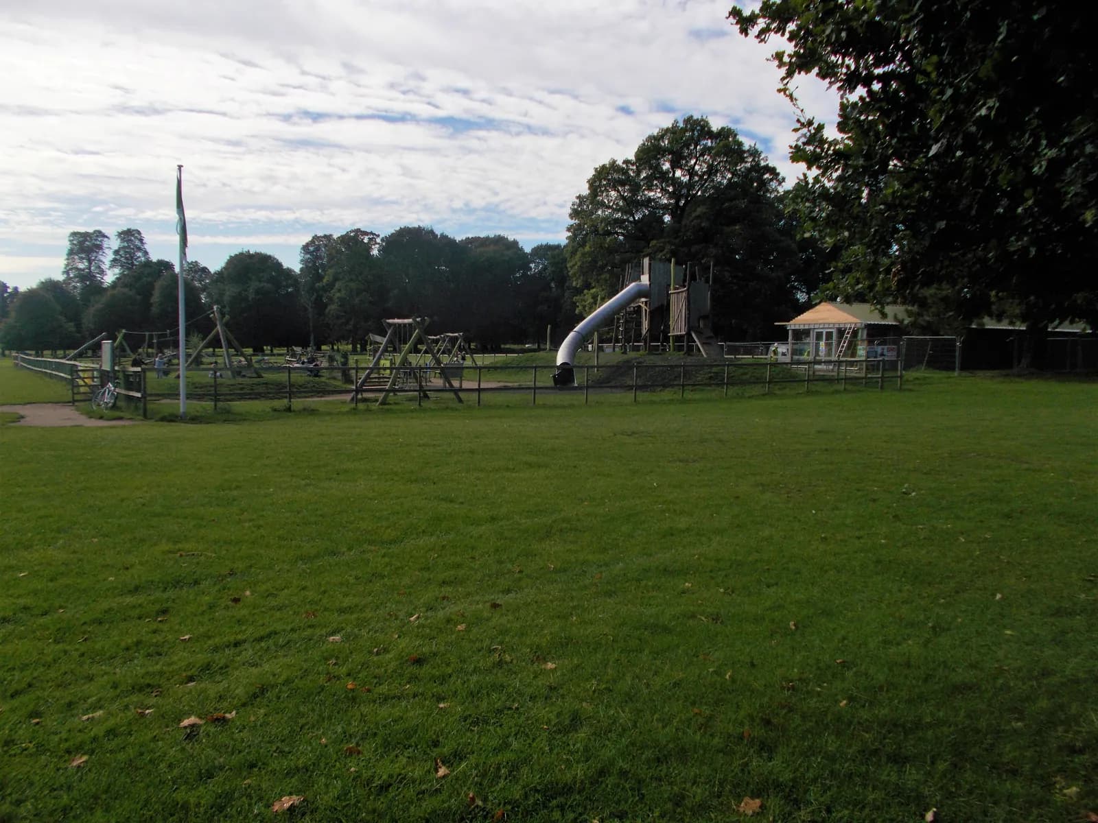 A wide shot of a playground with green grass in the foreground and various playground equipment, including swings and a slide, visible behind a fence, with trees and a building in the background under a cloudy sky.