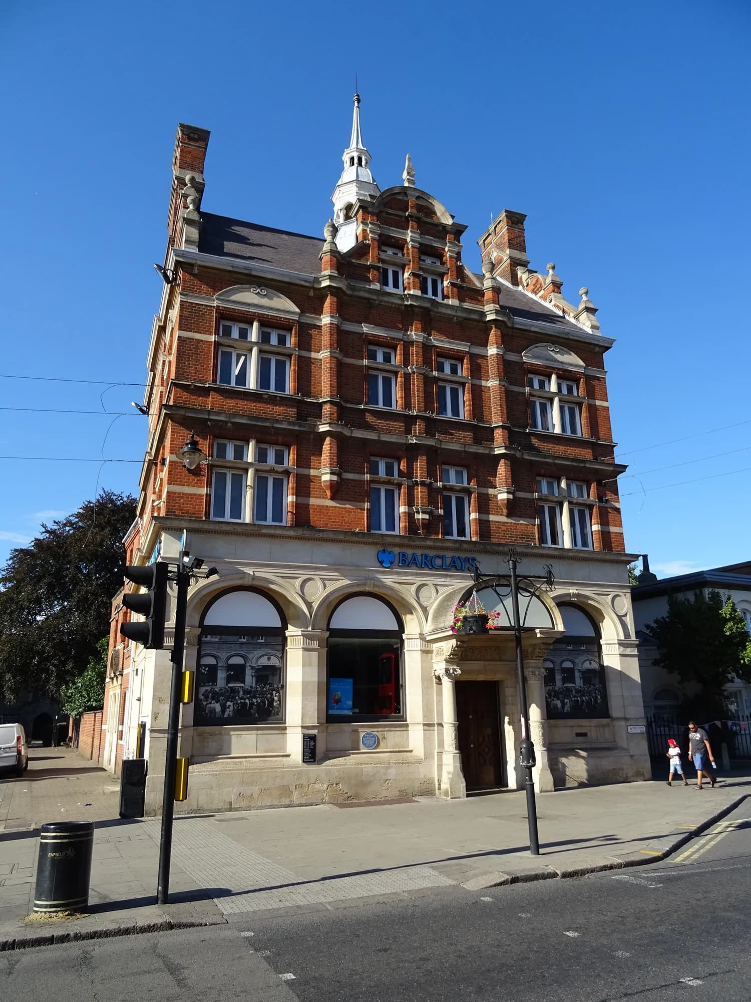 A grand, multi-story brick building with ornate architectural details and a steeple, housing a Barclays bank on the ground floor, stands under a clear blue sky, with a street and sidewalk in the foreground.