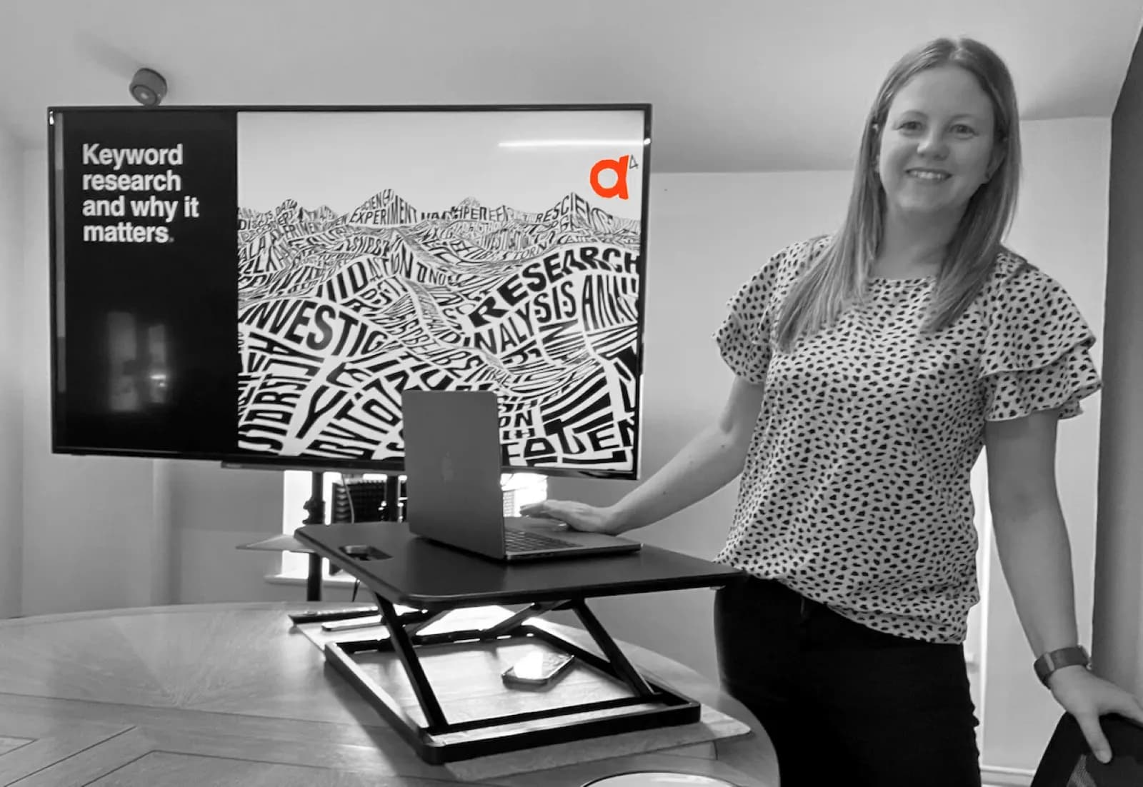 A smiling woman stands beside a desk with a laptop and a large screen displaying "Keyword research and why it matters" in black and white.