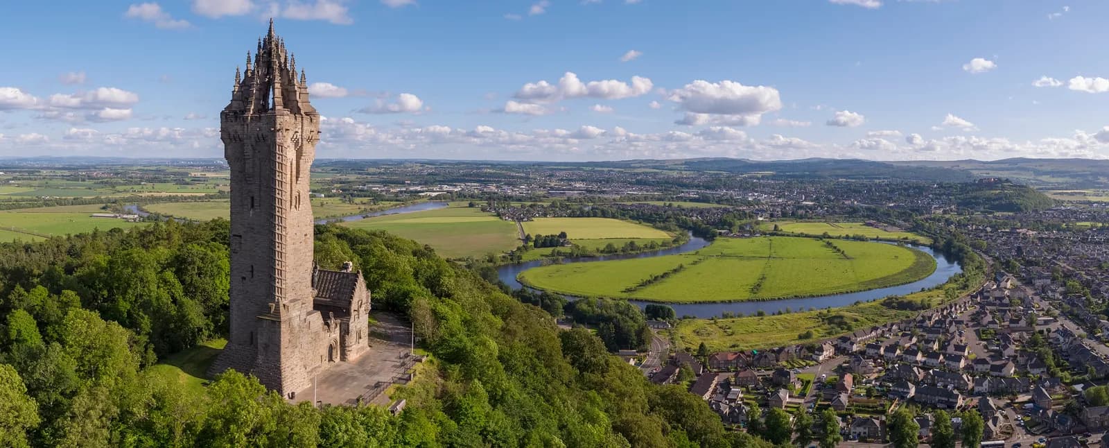 The National Wallace Monument stands tall on a lush green hill overlooking a meandering river, green fields, and a distant town under a cloudy blue sky.