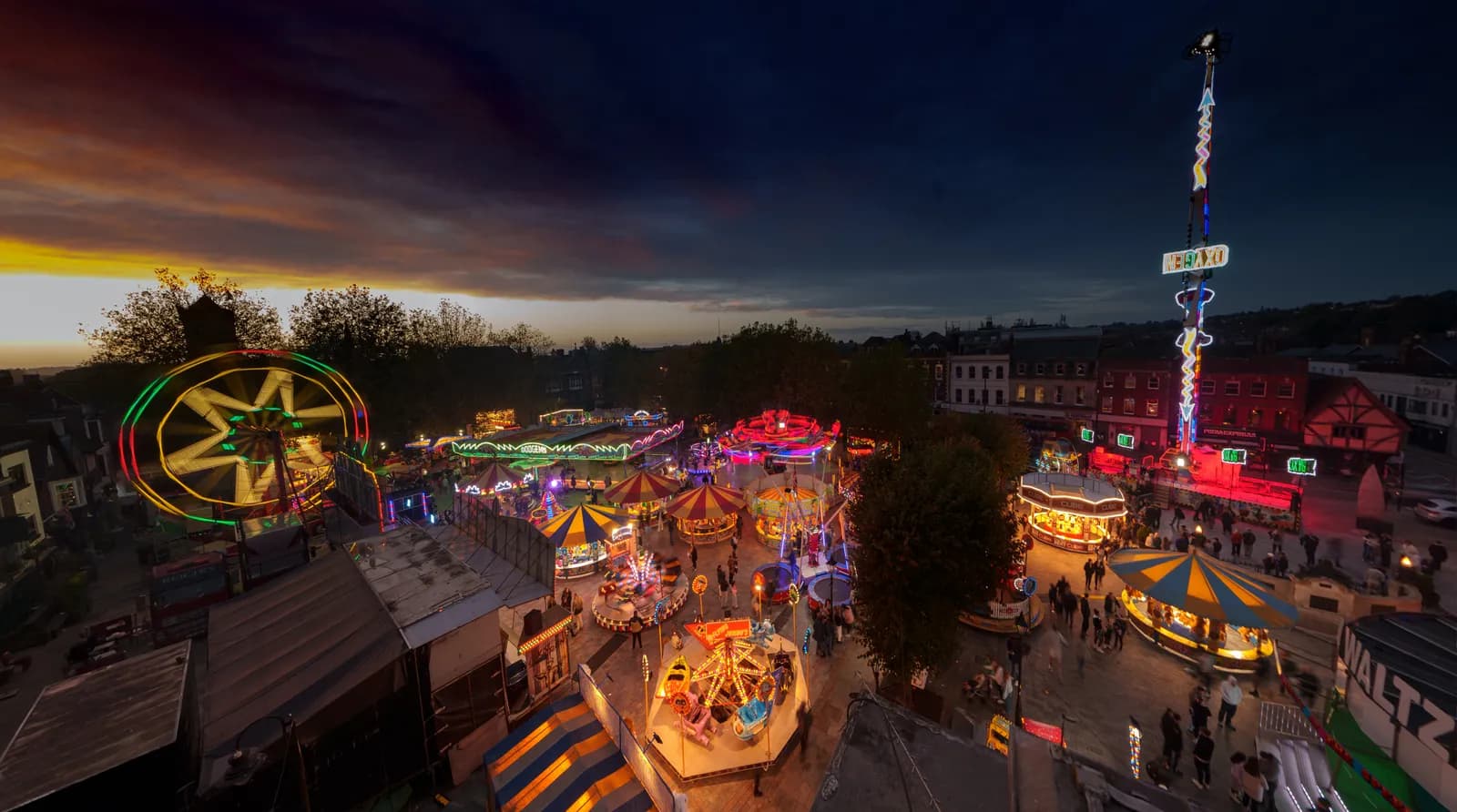 An aerial view of a vibrant funfair at dusk, featuring brightly lit rides including a Ferris wheel, carousels, and a tall drop tower, with people walking between attractions.