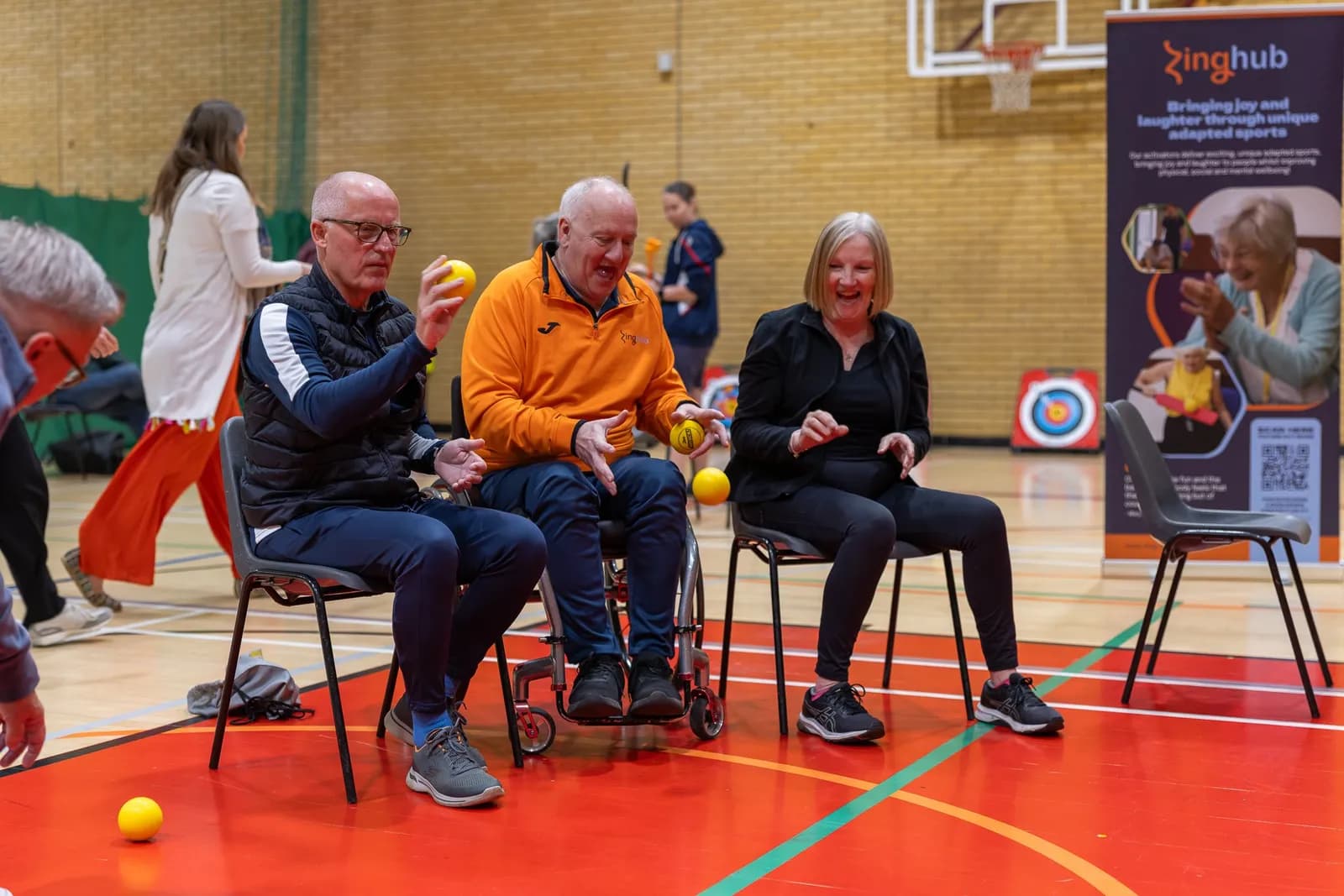 Three adults, two men and one woman, sit on chairs in a gym and happily play with small yellow balls, with a banner for 'Zinghub' in the background.