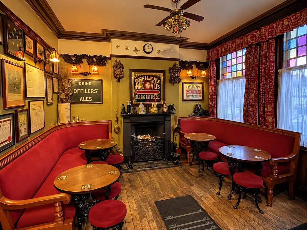 A cozy, traditional pub interior features red velvet banquettes and stools, small wooden tables, framed pictures on the wall, a fireplace, and stained-glass windows with red patterned curtains.