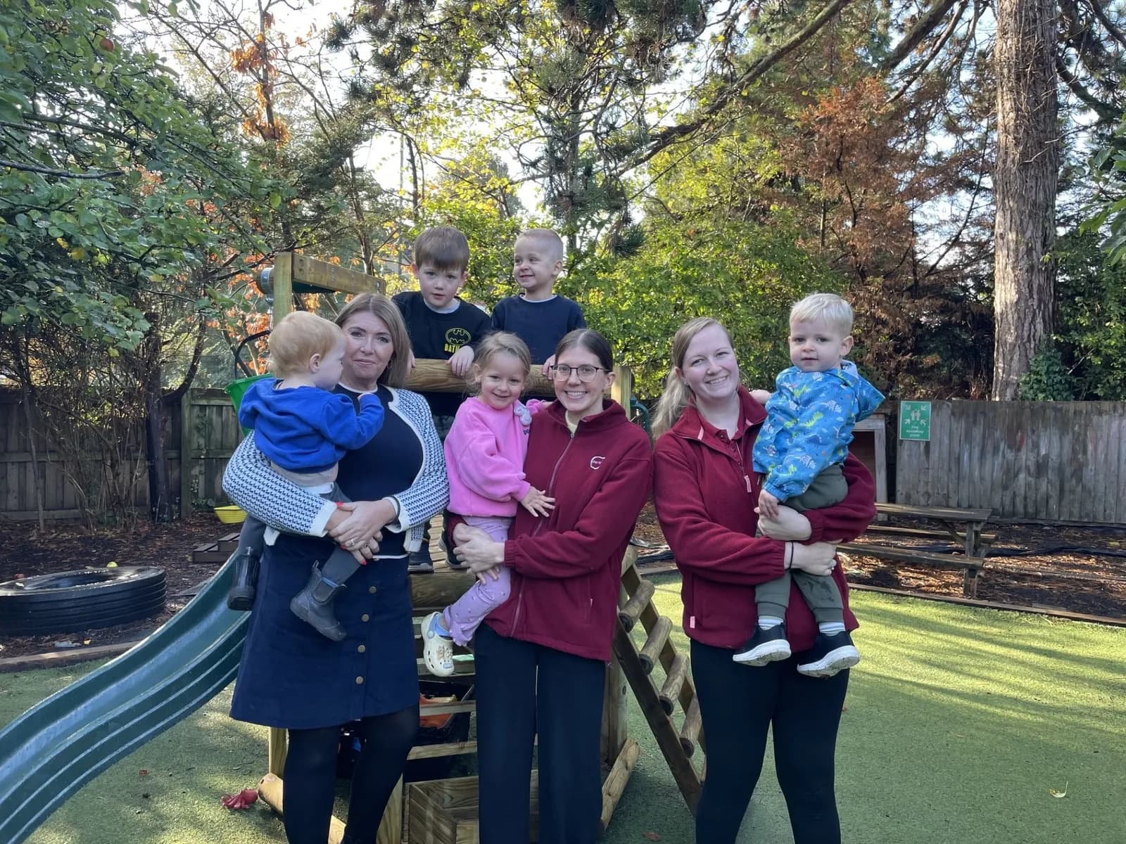 Four adults and five children pose for a picture outdoors near playground equipment and trees.