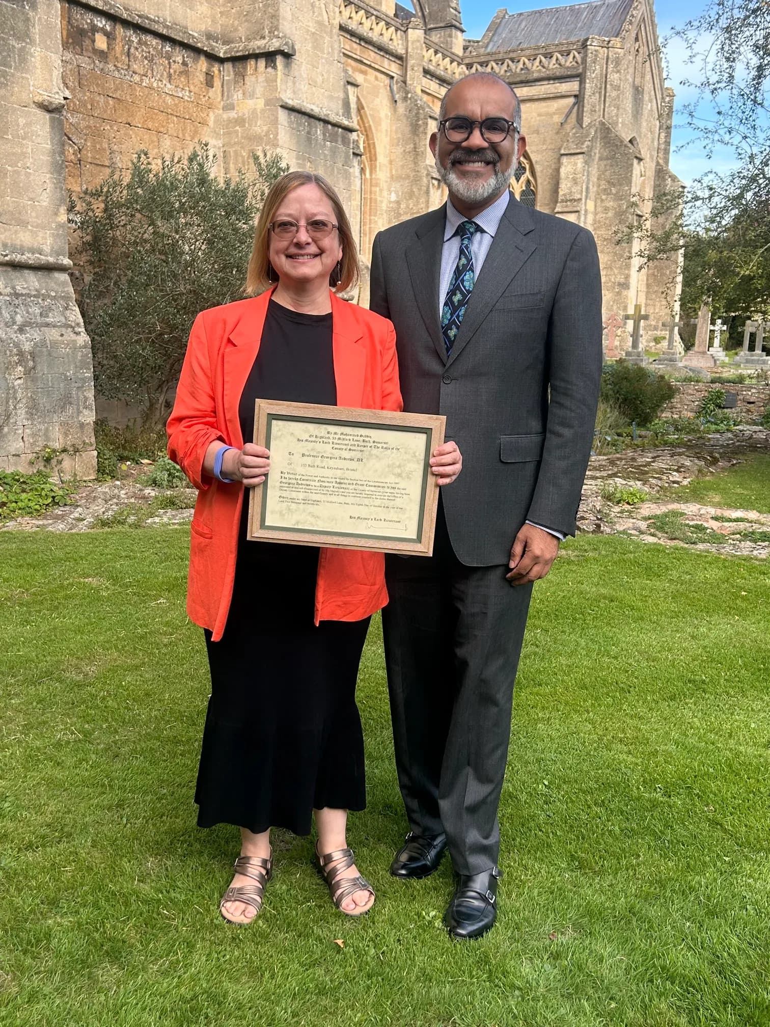 A woman in an orange blazer and a man in a grey suit stand on a grassy lawn with an old stone building in the background, the woman holding a framed certificate.