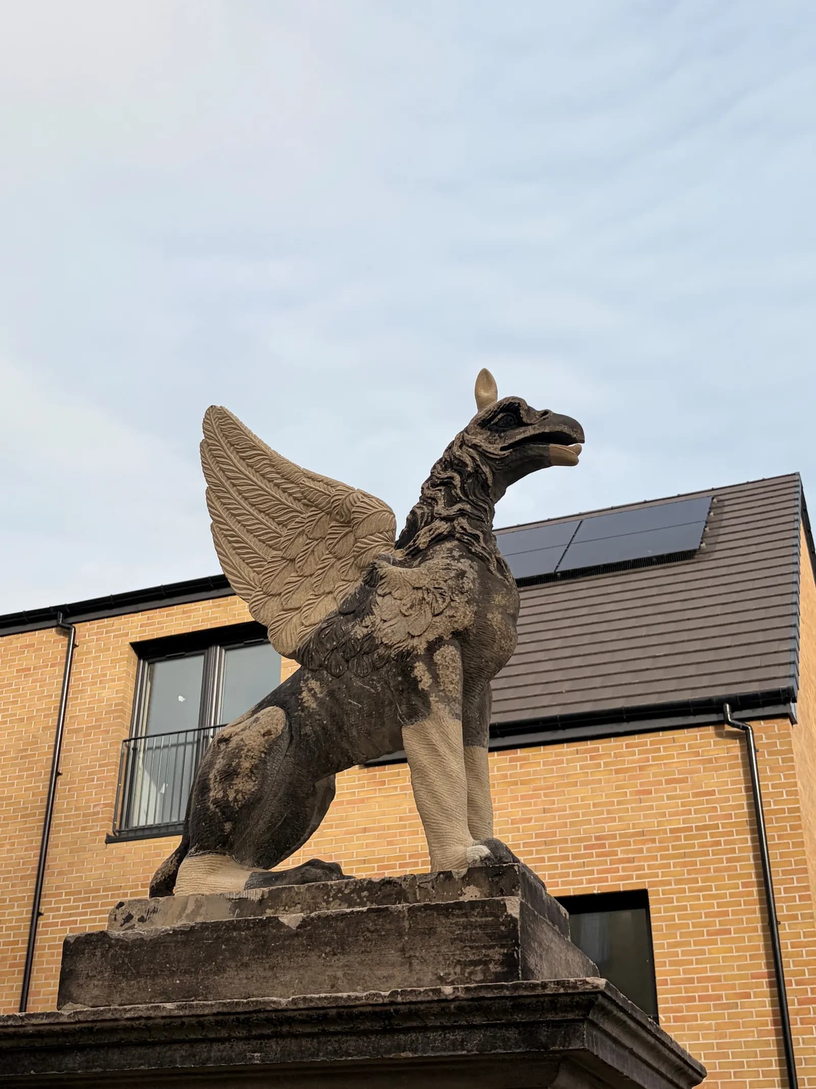 A weathered stone griffin statue with intricate wing details stands on a pedestal, with a modern brick building and a gray sky in the background.