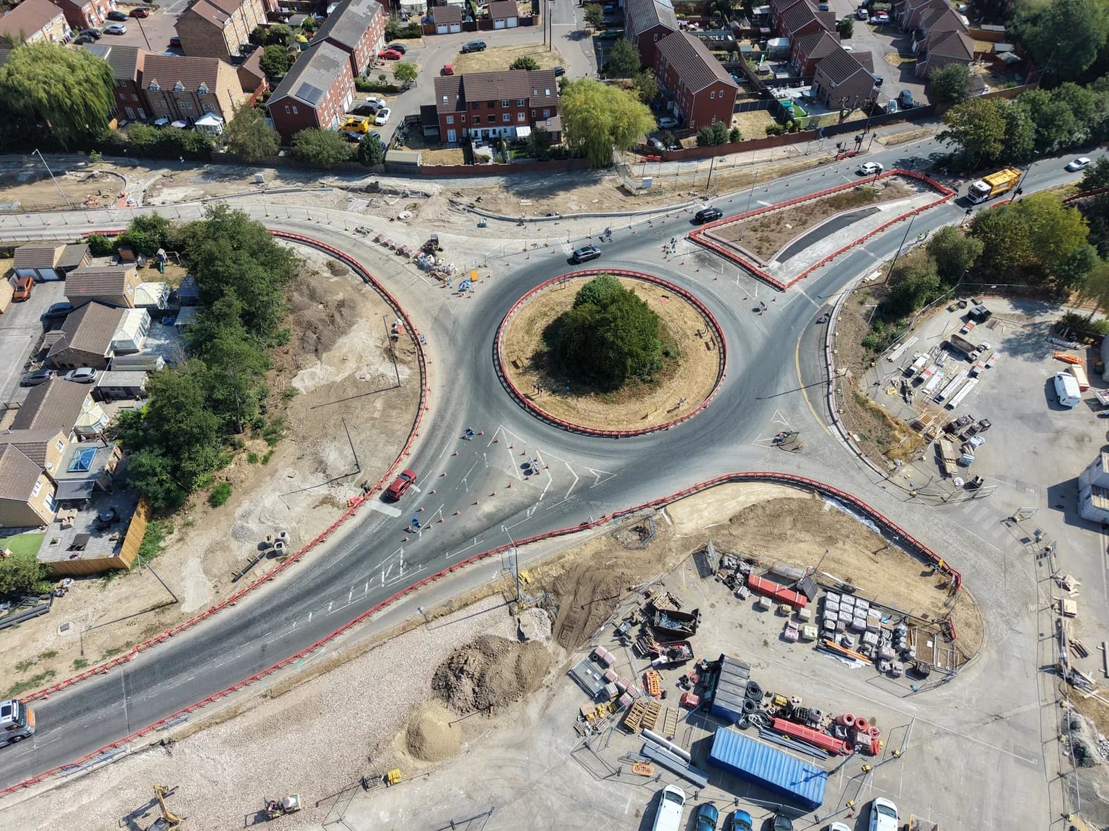 An aerial view captures a newly constructed roundabout with an island of green trees at its center, surrounded by roads leading to residential areas and a construction site.