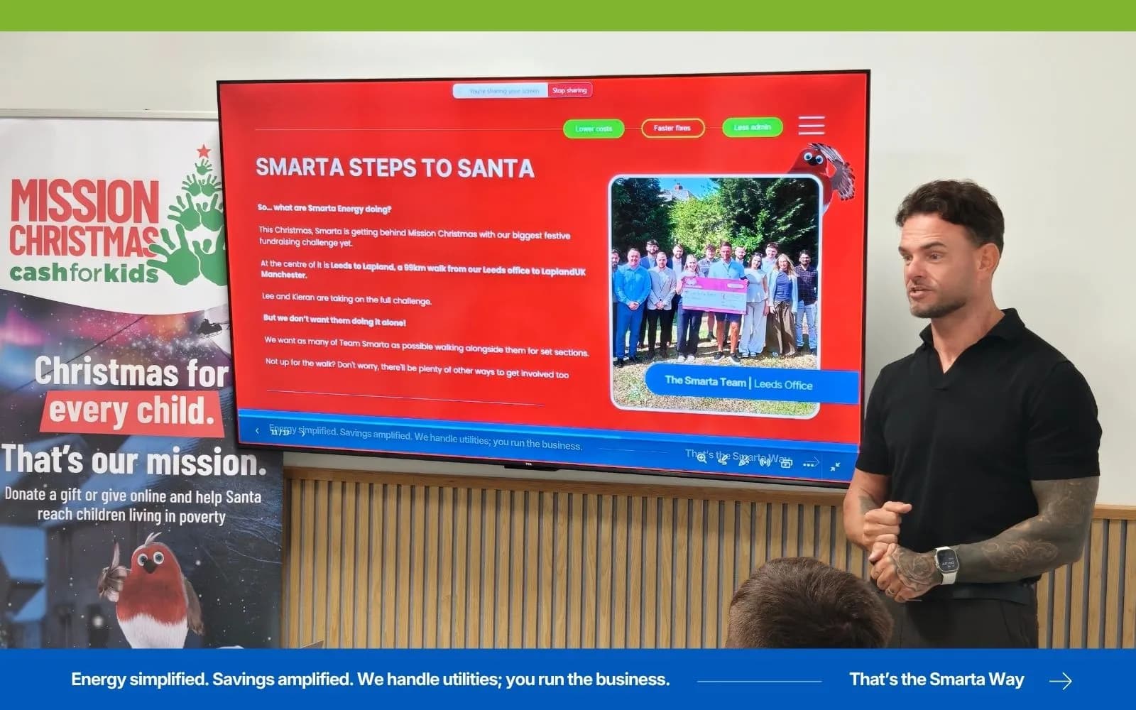 A man in a black polo shirt and tattooed arm is presenting in front of a large screen displaying information about "Smarta Steps to Santa" and a team photo.