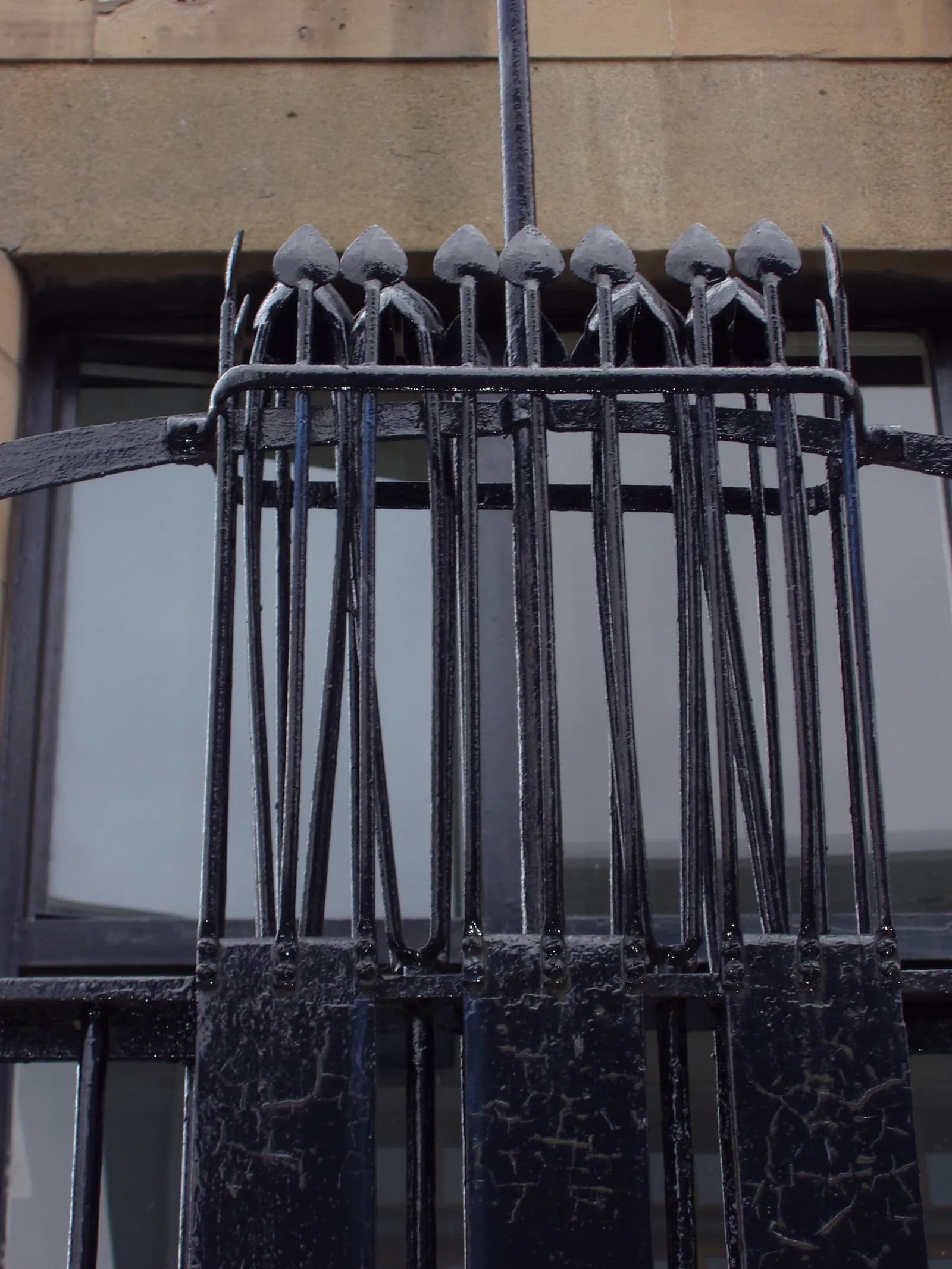 A close-up view of a dark metal ornate railing with pointed finials against a blurred background of a building facade and a window.