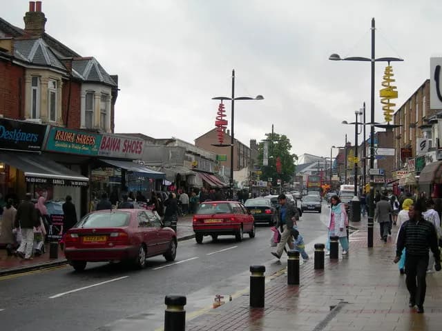 A busy street scene with cars driving on a wet road, pedestrians walking on sidewalks, and shops lining the left side of the street under an overcast sky.