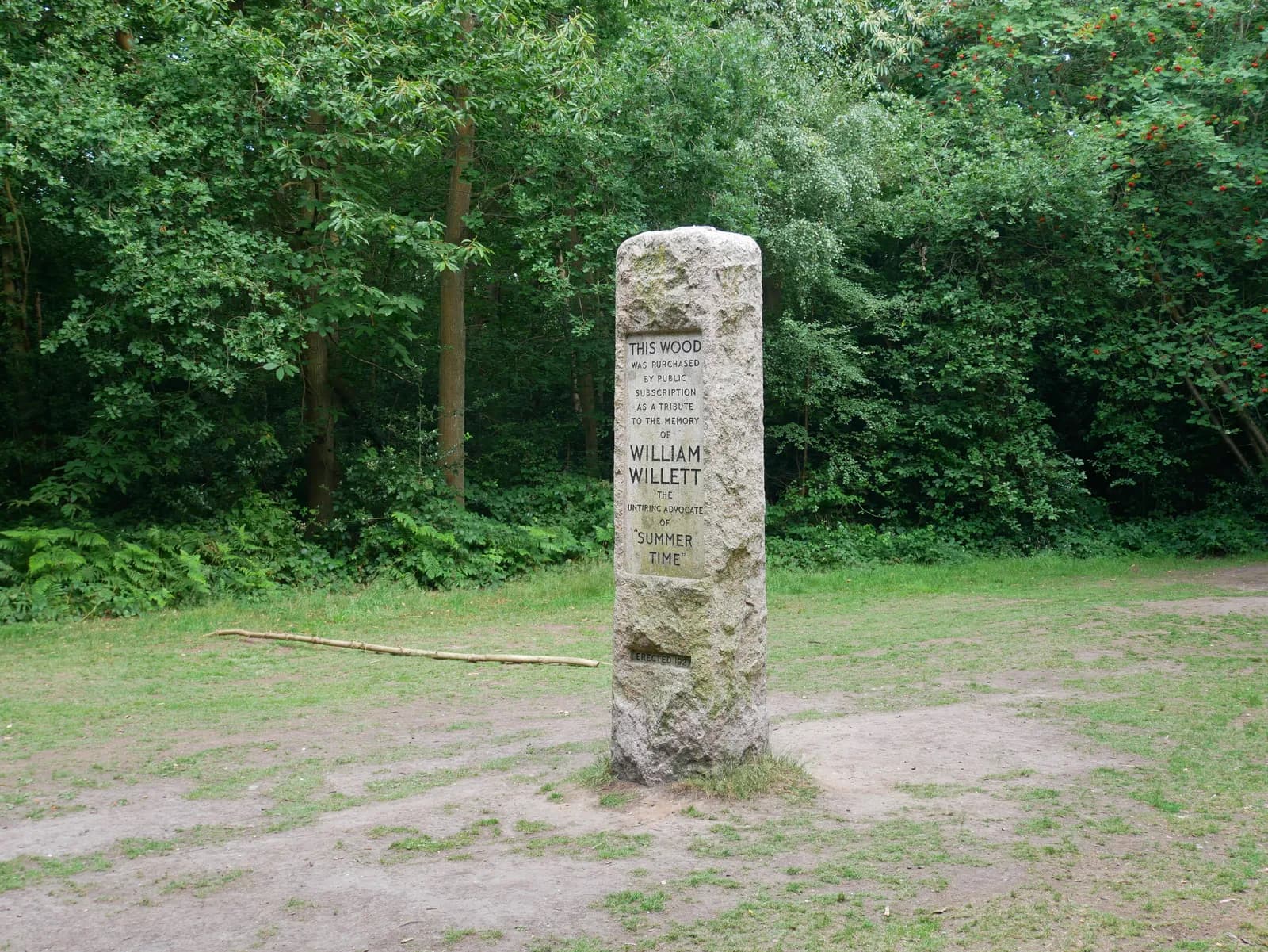 A stone monument stands tall in a wooded area, with text honoring William Willett and mentioning summer time, surrounded by green foliage and a dirt path.