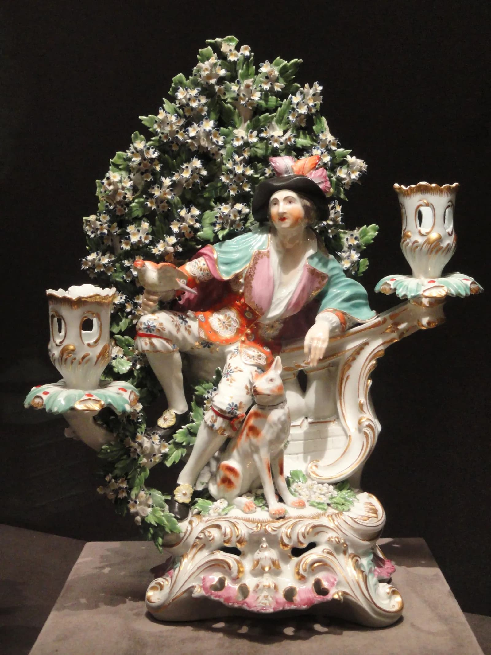 A Rococo-style porcelain candelabra features a male figure in 18th-century attire holding a bird, with a dog at his feet, all set against a backdrop of flowering foliage and ornate white and gold candleholders.