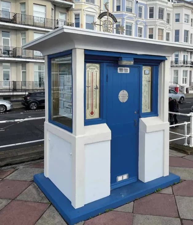 A small blue and white kiosk with a weather vane on its roof stands on a paved sidewalk next to a road, with multi-story buildings in the background.