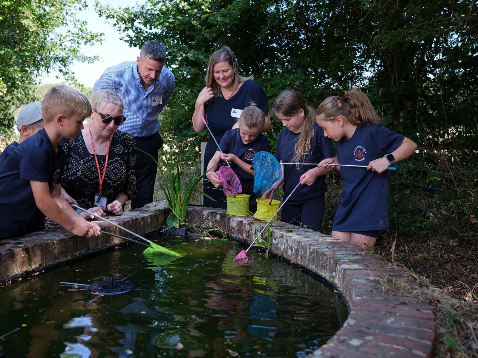 A group of adults and children are gathered around a brick-lined pond with dark water, using small nets on poles to catch things in the water.