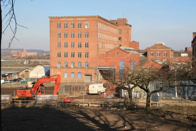 A large red brick mill building with many windows stands prominently against a clear sky, with an orange excavator and construction site in the foreground.