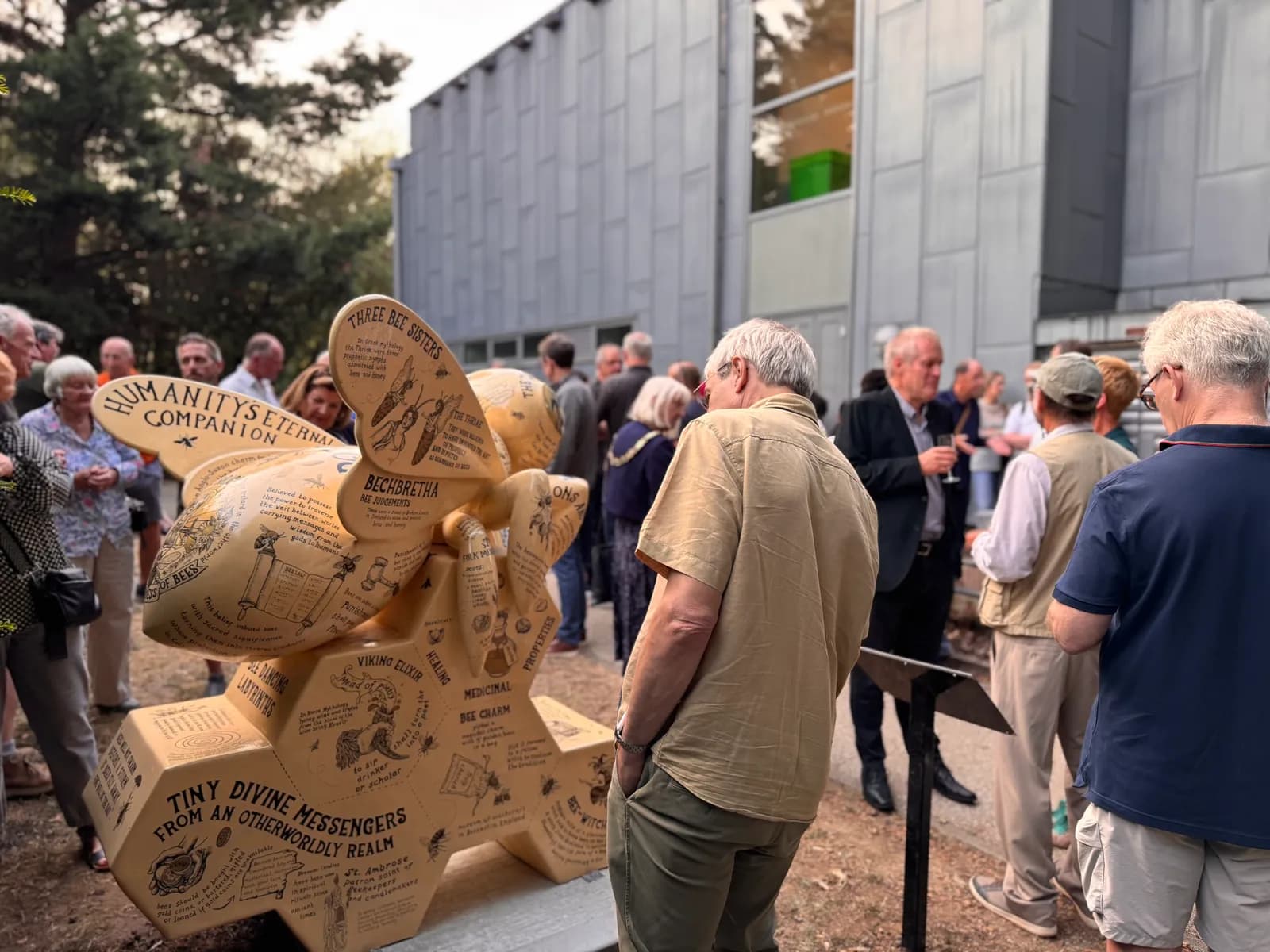 A honeybee sculpture with text, on display outdoors with people admiring it and a building in the background.