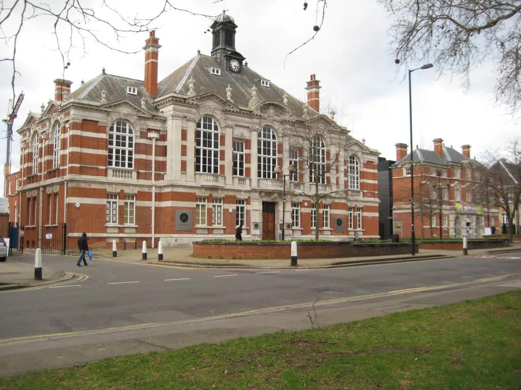 A historic red brick and white classical style building with a clock tower and multiple arched windows stands next to a street, with residential buildings and trees in the background.