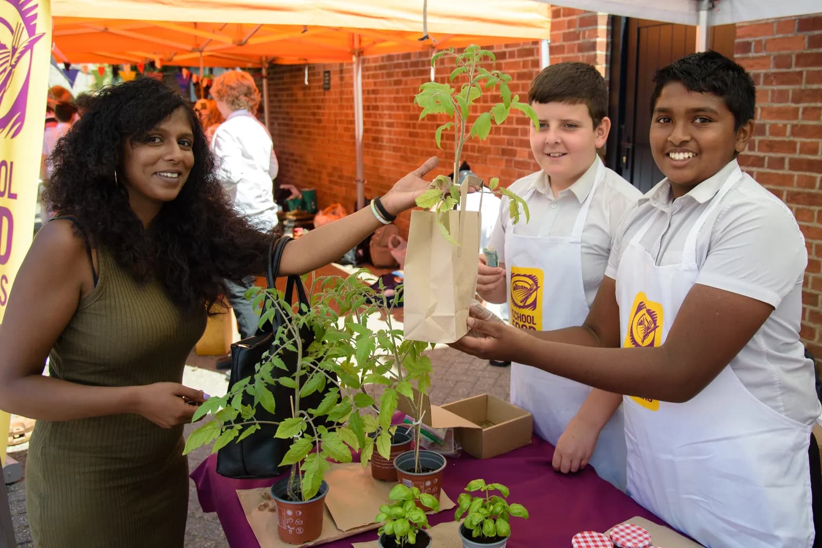 A smiling woman with dark curly hair hands a potted plant in a paper bag to a smiling boy wearing a white apron, next to another similarly dressed boy, at a market stall.