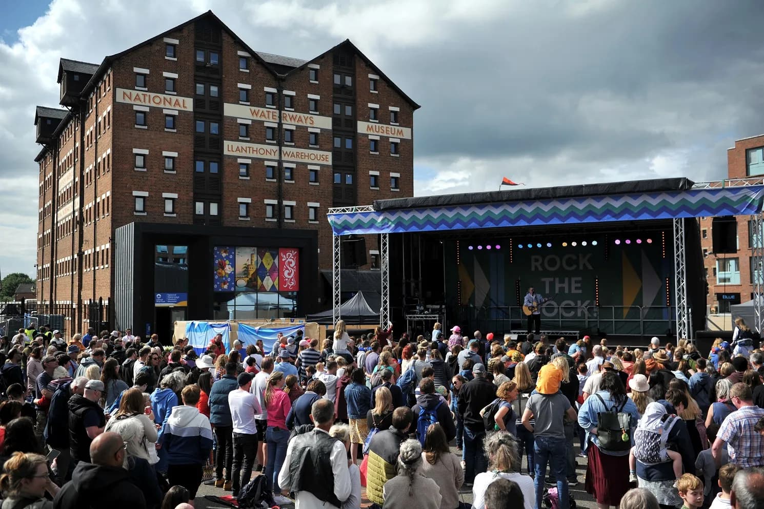 A large crowd of people gathered in front of a stage where a person is performing, with a red brick building labeled "National Waterways Museum Llanthony Warehouse" in the background under a cloudy sky.