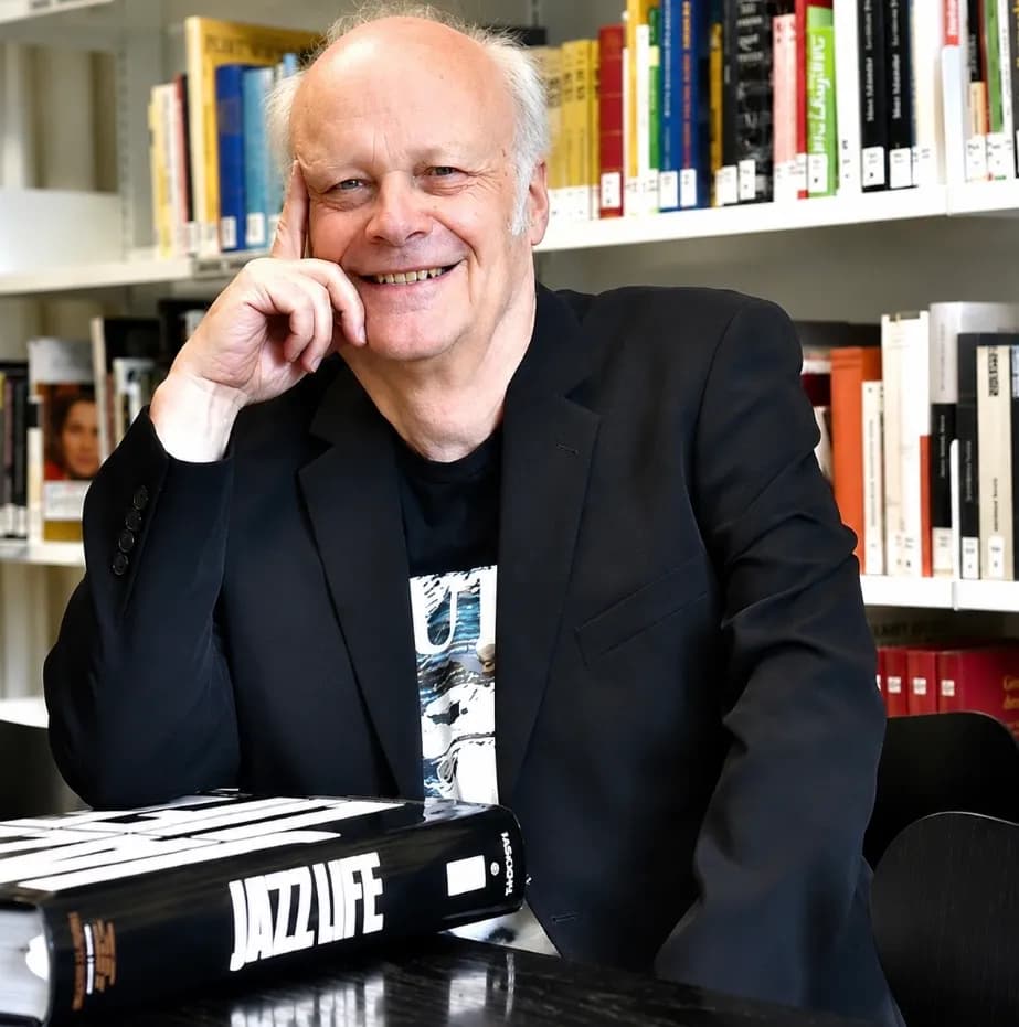 A smiling man with light hair and a black jacket rests his hand on his chin, sitting at a table with a book titled "JAZZ LIFE" in front of him, with bookshelves in the background.