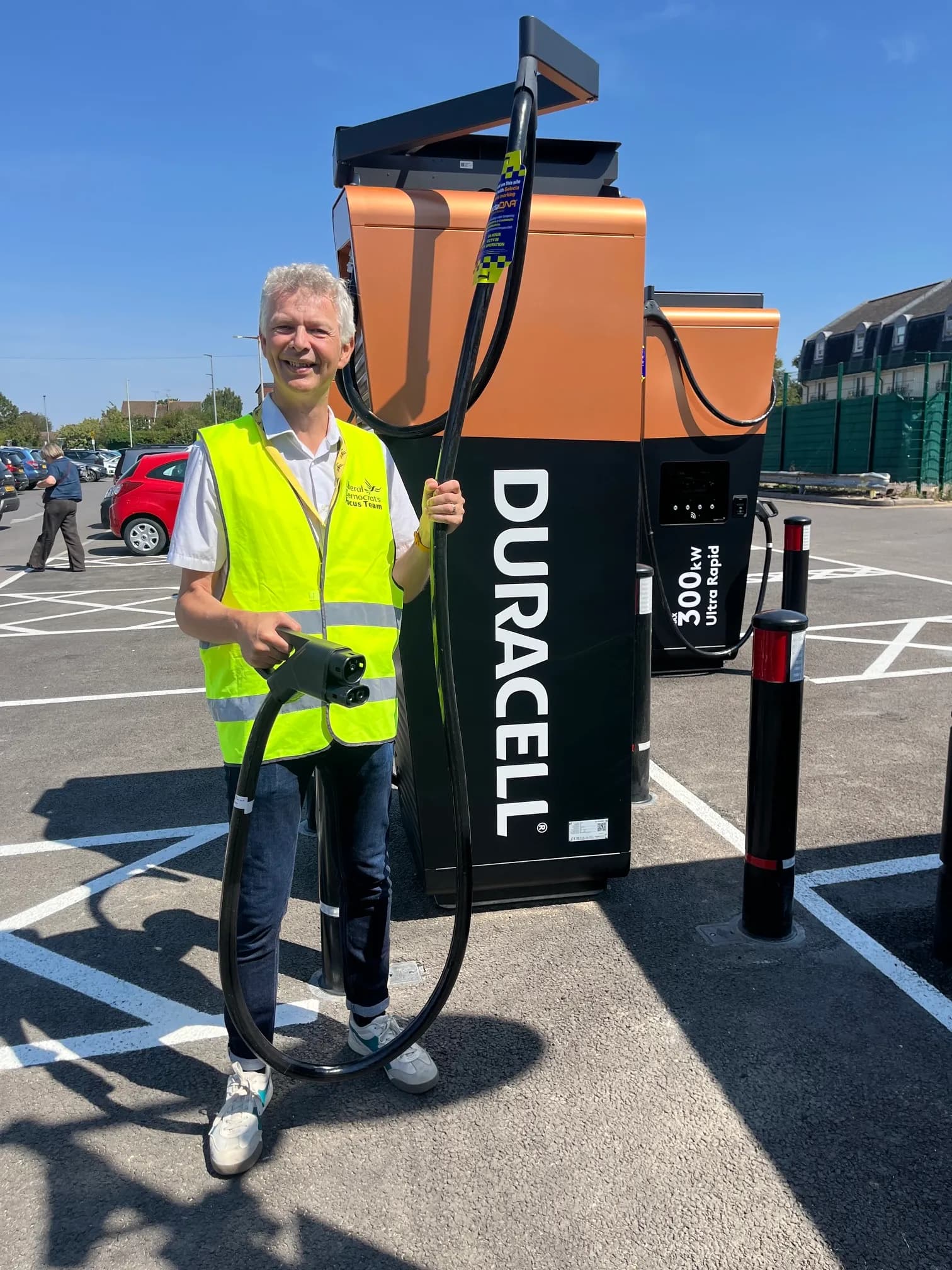 A man in a high-visibility vest stands proudly next to a large Duracell-branded electric vehicle charging station, holding the charging cable.