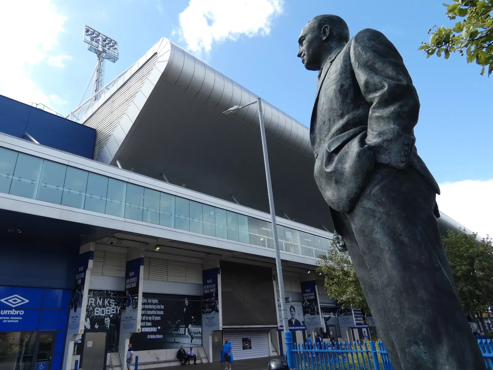 A bronze statue of a man in a suit, seen from a low angle, stands in front of a modern stadium with a blue and metallic facade under a partly cloudy sky.