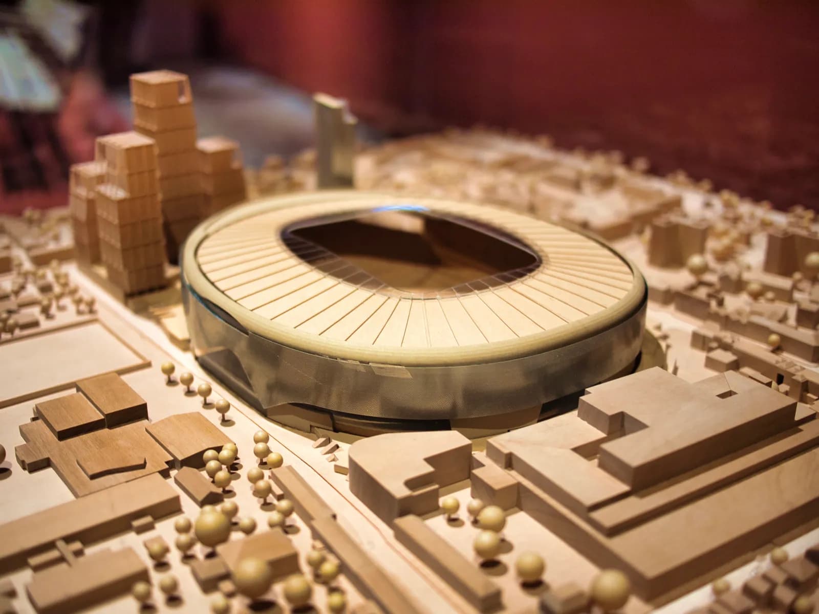 A detailed architectural model showcases a modern stadium with a light-colored, slatted roof, surrounded by numerous smaller buildings and miniature trees on a light brown base.