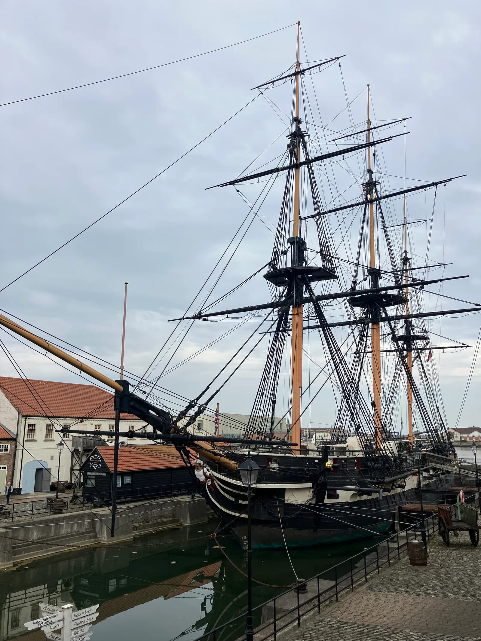 A large mast ship with black and white hull details is docked in a dry dock, surrounded by historic buildings under a cloudy sky.