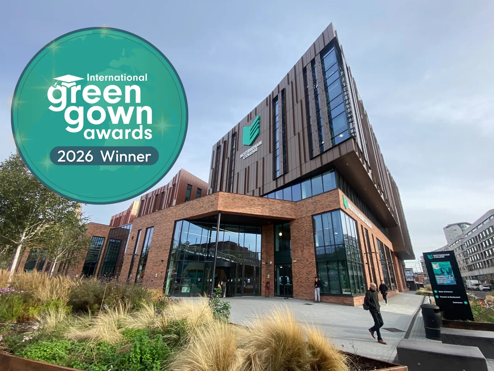 A modern building with large windows and brick accents, featuring an "International Green Gown Awards 2026 Winner" overlay in the upper left corner, with landscaping in the foreground and people walking near the entrance.