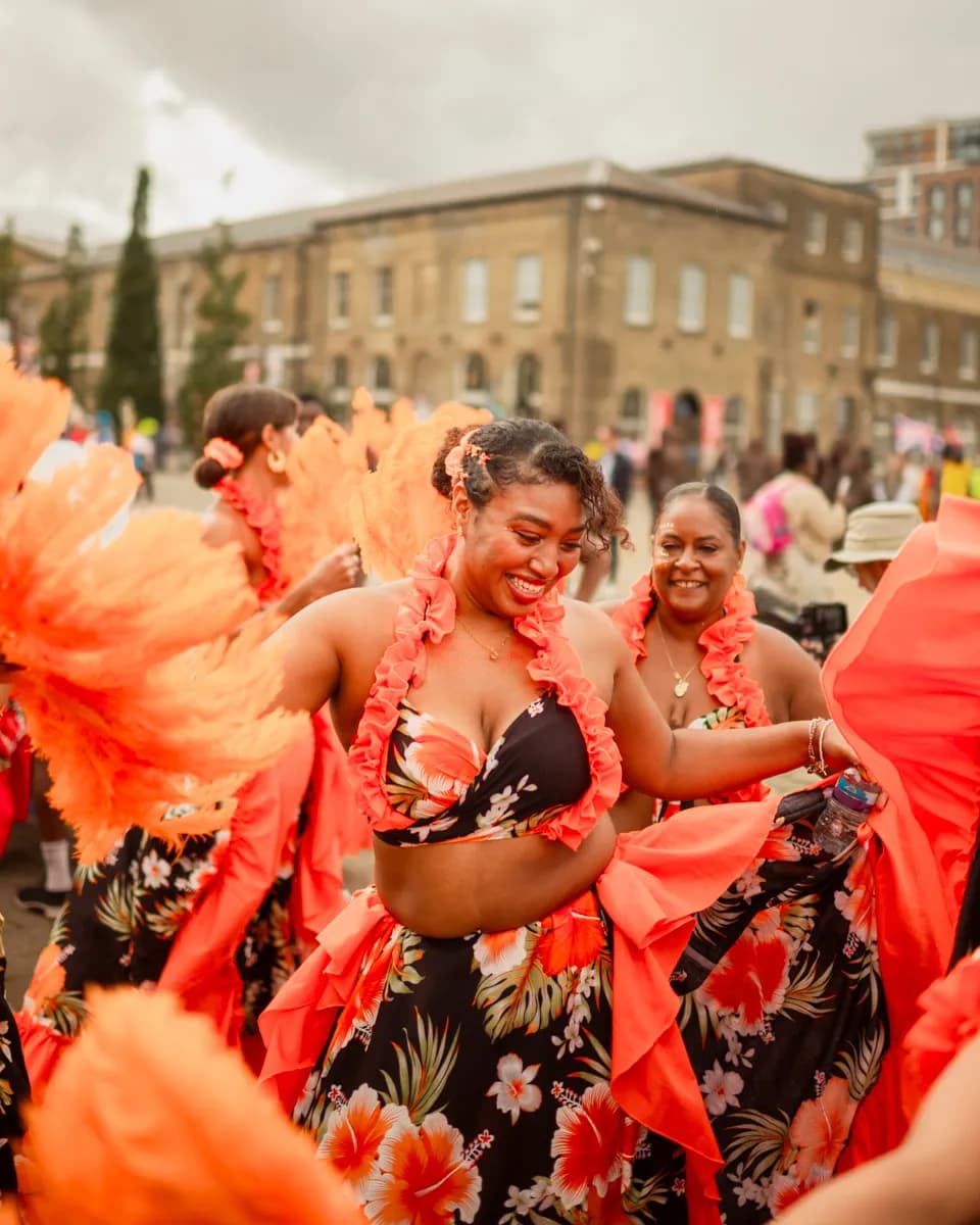 A woman in a floral carnival outfit with orange ruffles and a flower necklace smiles broadly while dancing, surrounded by other performers and a building in the background.