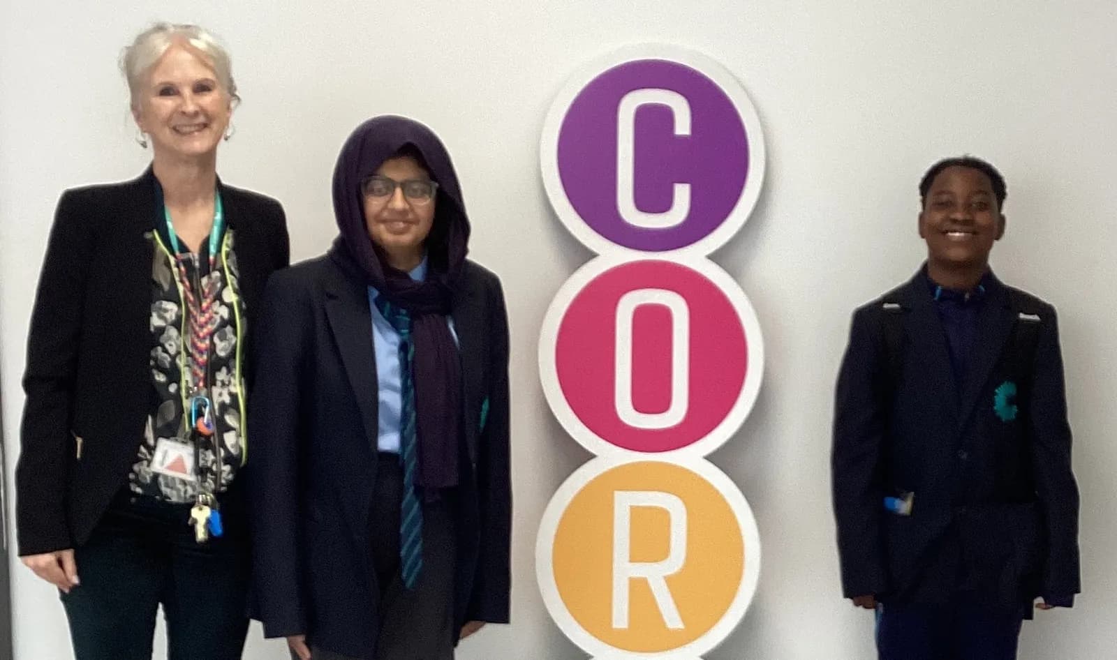 A woman with gray hair and two students in school uniforms stand smiling in front of a white wall with vertical stacked colorful circles spelling 'COR'.