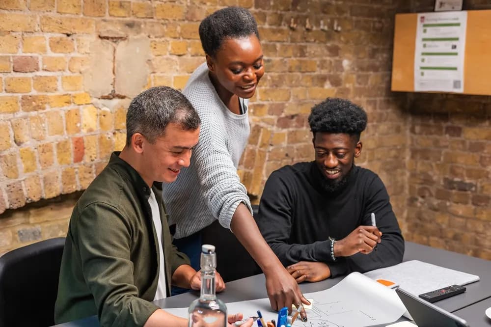 Three people, two men and one woman, are gathered around a table in a brick-walled room, looking at a document and smiling, with the woman pointing at something on the paper.