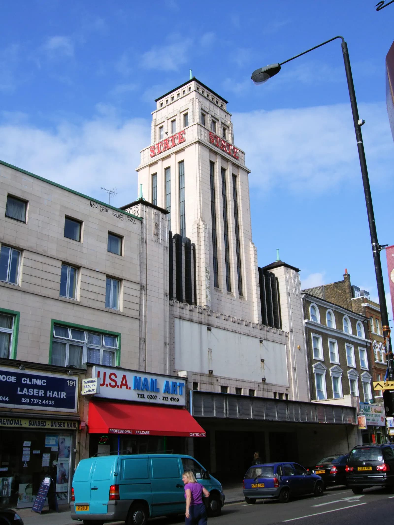 A tall, white art deco building with red 'STATE' signs dominates the center of a bustling street scene, flanked by lower commercial buildings and several cars.