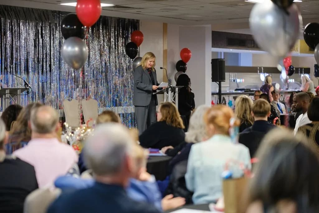 A woman in a gray suit stands at a podium giving a speech to a seated audience at an event with red, black, and silver balloons.