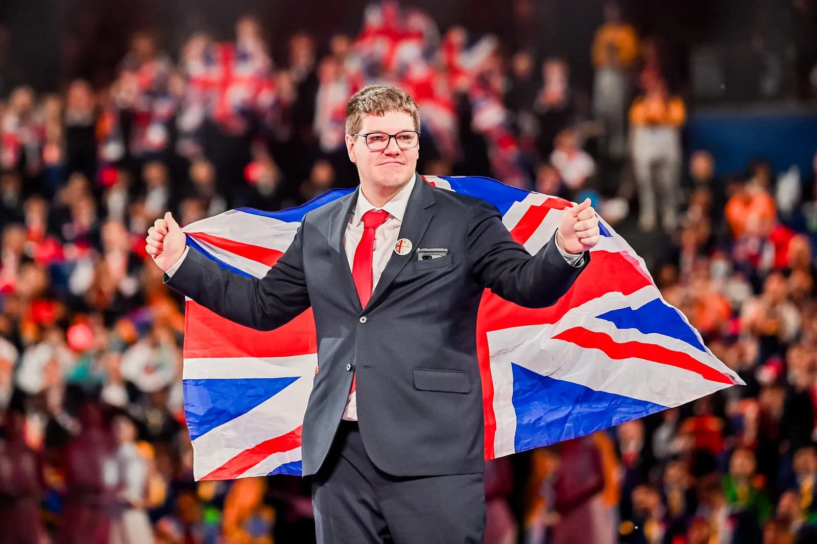 A man in a suit and red tie proudly holds a British flag behind him, with a blurred crowd in the background.