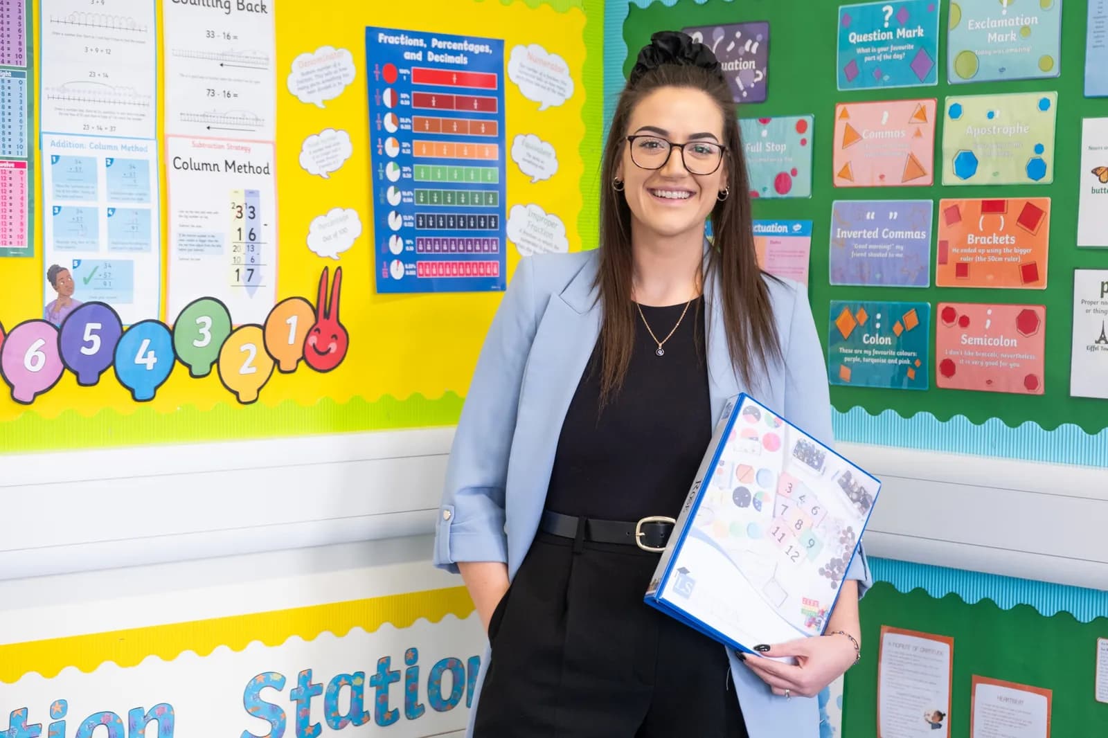 A smiling female teacher with dark hair and glasses stands in a classroom, holding a folder, with colorful educational charts on the wall behind her.