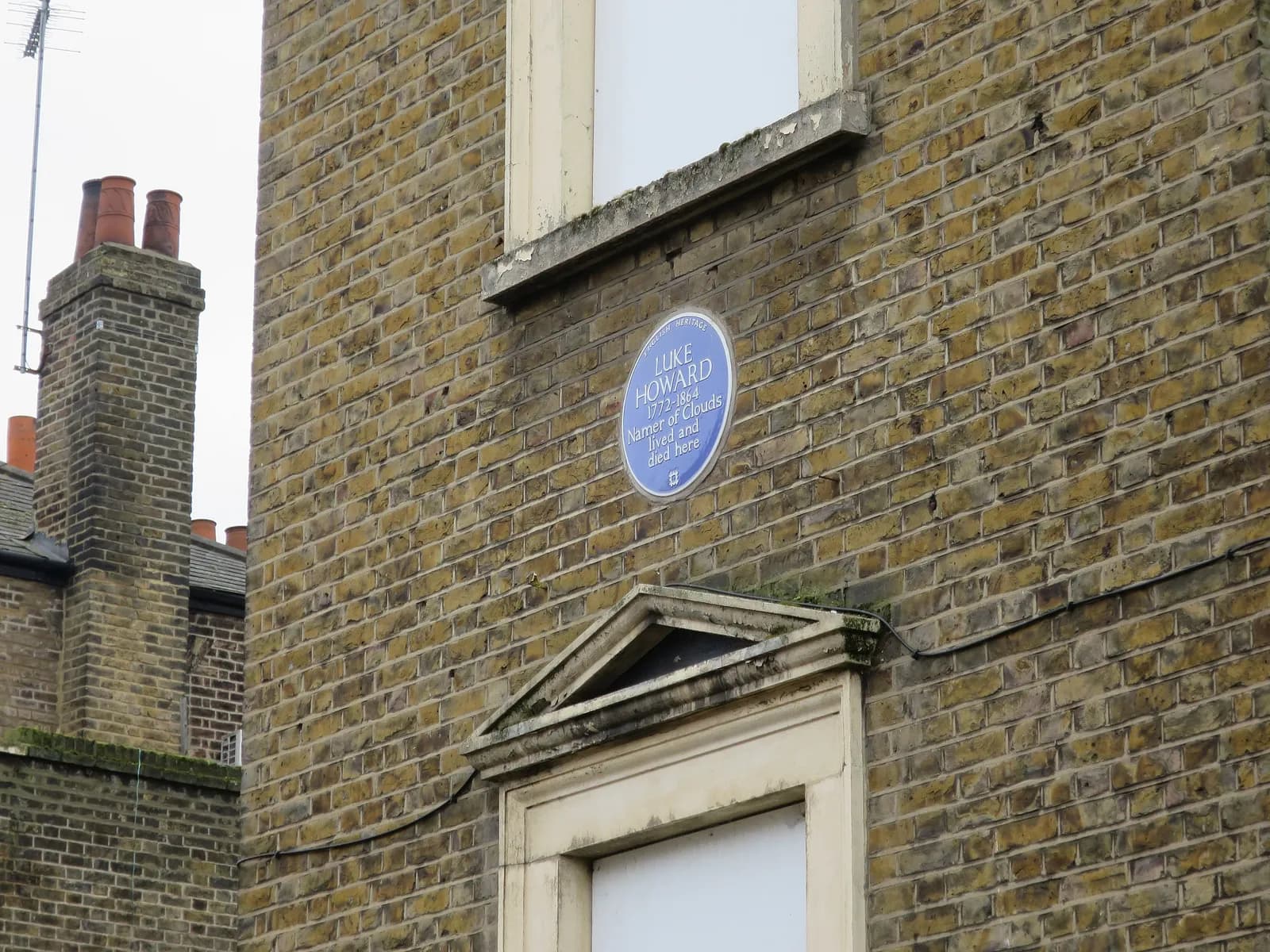 A blue historical plaque for Luke Howard is affixed to a textured brick wall of a building, with a window and architectural detailing beneath it, and a chimney structure to the left.