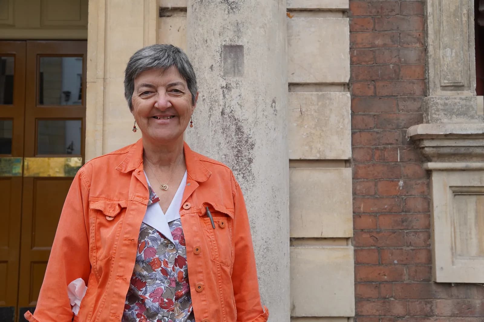 A smiling woman with short gray hair and an orange jacket stands in front of a building with brick and stone elements.