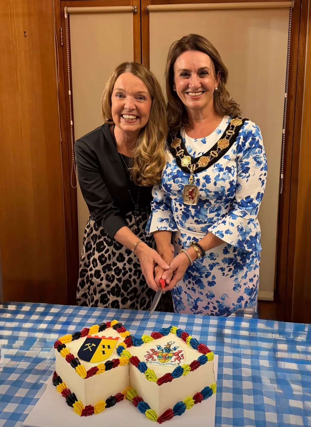 Two smiling women, one in a blue and white floral dress with a mayoral chain, jointly cut a decorated cake with two distinct crests on top, sitting on a blue and white checkered tablecloth.