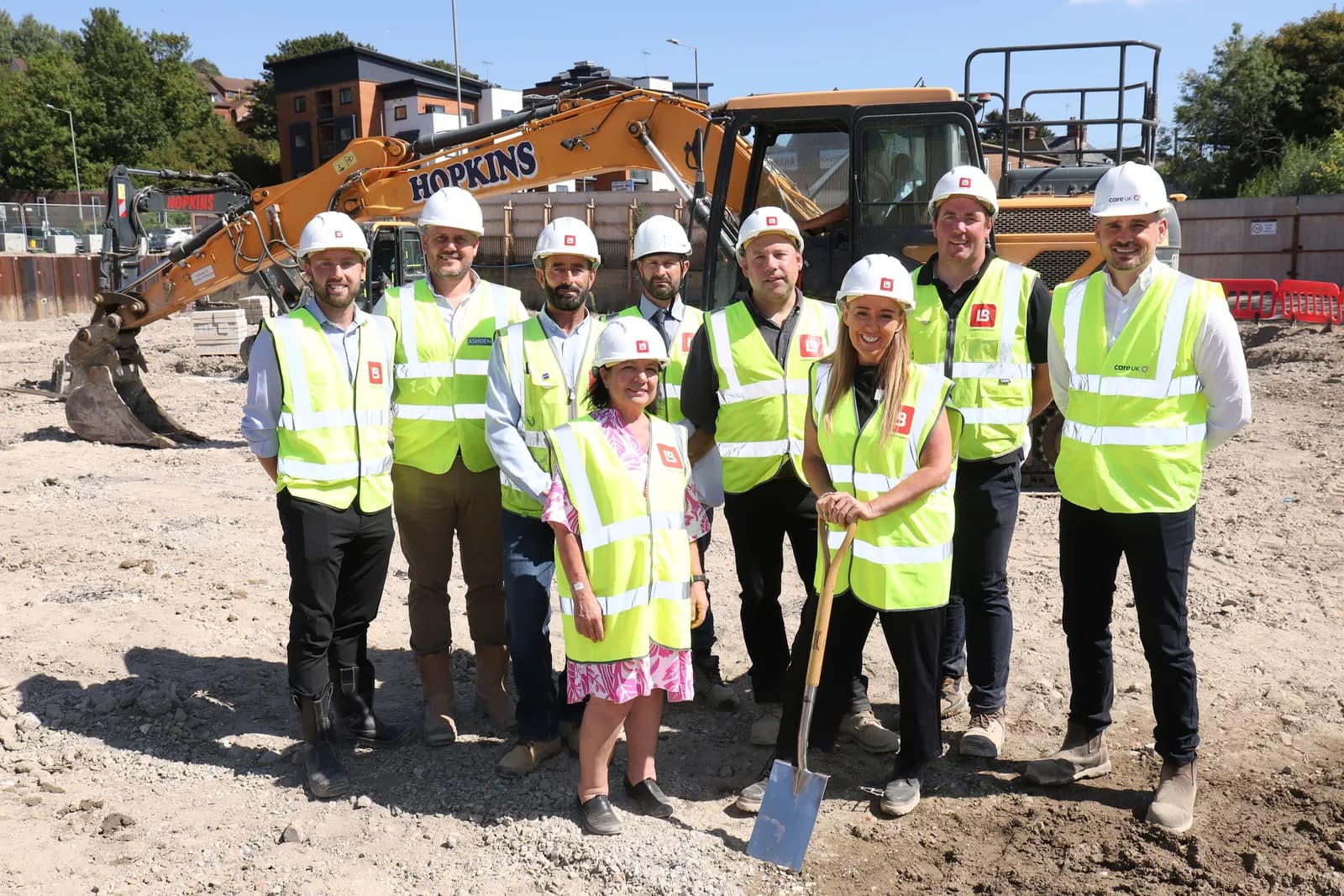 A group of nine people in construction hard hats and reflective vests stand on a dirt lot in front of a large yellow excavator.