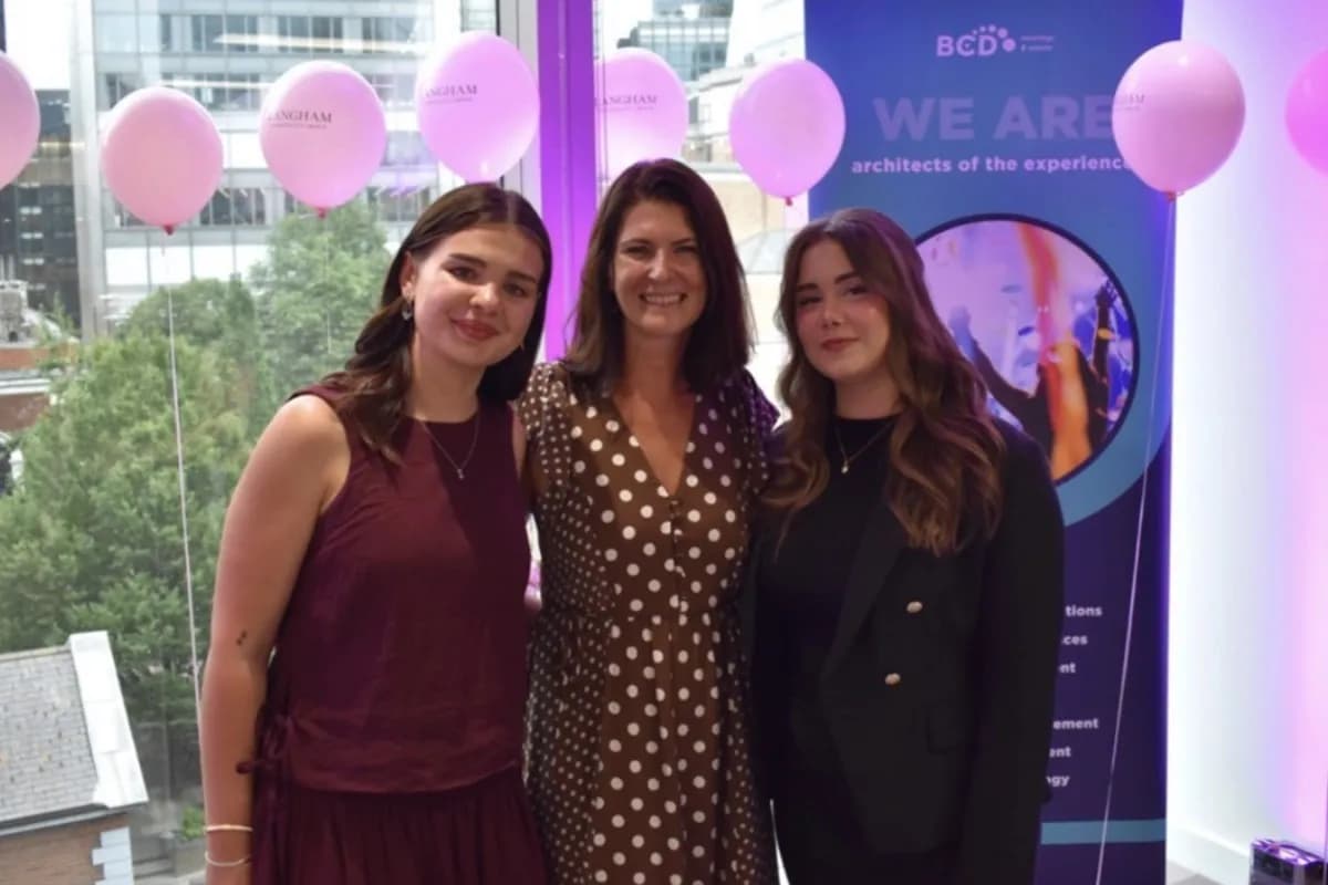 Three women smile while standing in a room decorated with pink balloons and a banner for 'BCD: We Are Architects of the Experience'.
